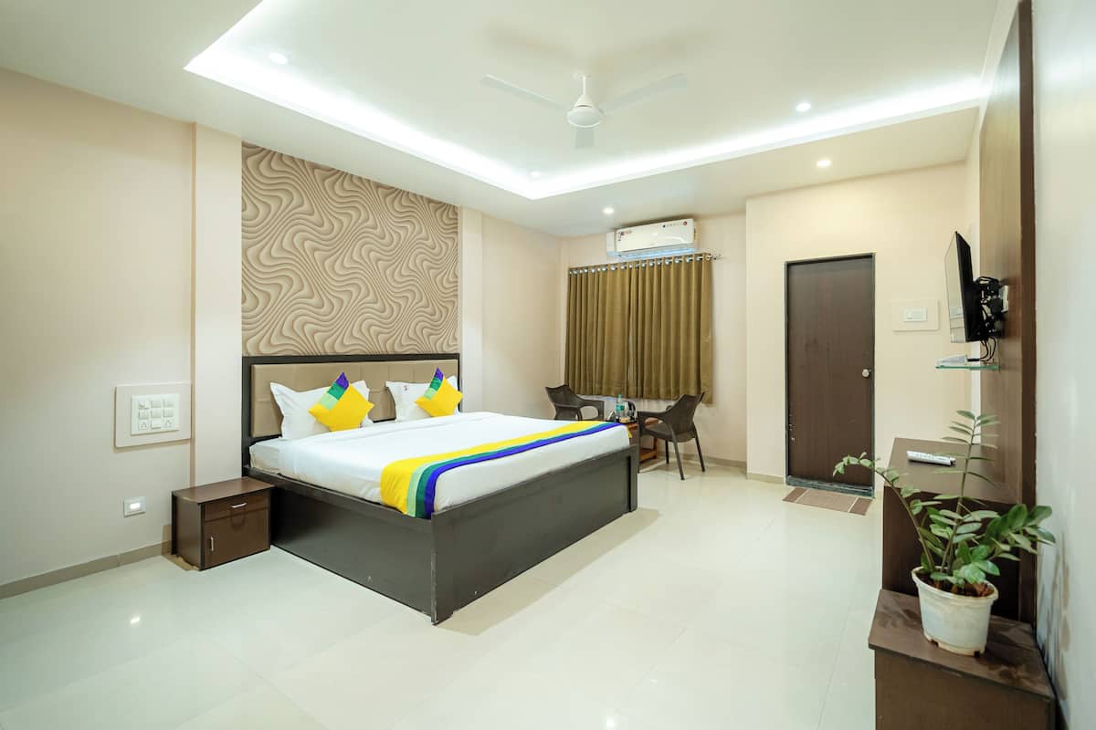 A modern hotel room with a large bed, bedside tables, a desk with chairs, a TV, and a potted plant.