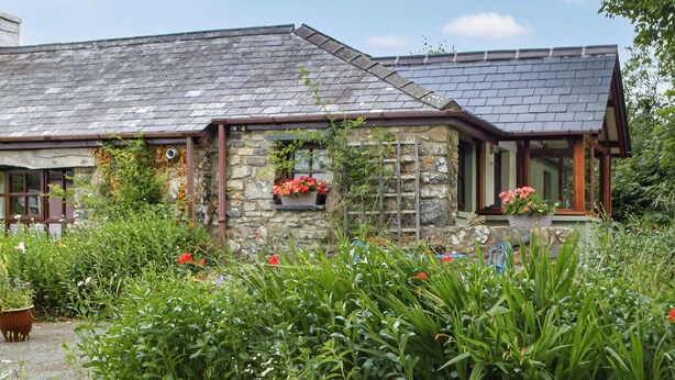 A stone house with a sloped roof, stone walls, and a garden with various plants and flowers.