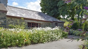 A stone building with a sloped roof, surrounded by a garden with white and purple flowers.