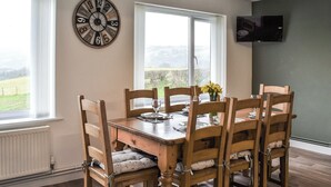 A dining room with a wooden table and chairs, a large wall clock, and a television mounted on the wall. - 4 bedroom accommodation in Corwen (Corwen)