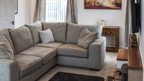 A living room with a grey sectional sofa, a black rug, a wooden side table, a painting of a forest scene, and a television on a stand. - 4 bedroom accommodation in Corwen (Corwen)