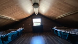 Quarto com teto inclinado, duas camas com roupa de cama azul, uma pequena janela e piso de madeira.