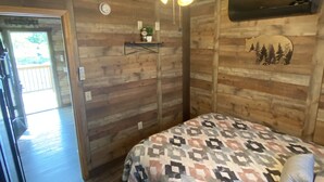 Quarto de cabana de madeira com cama, ventilador de teto, prateleira na parede e porta que dá para área externa.