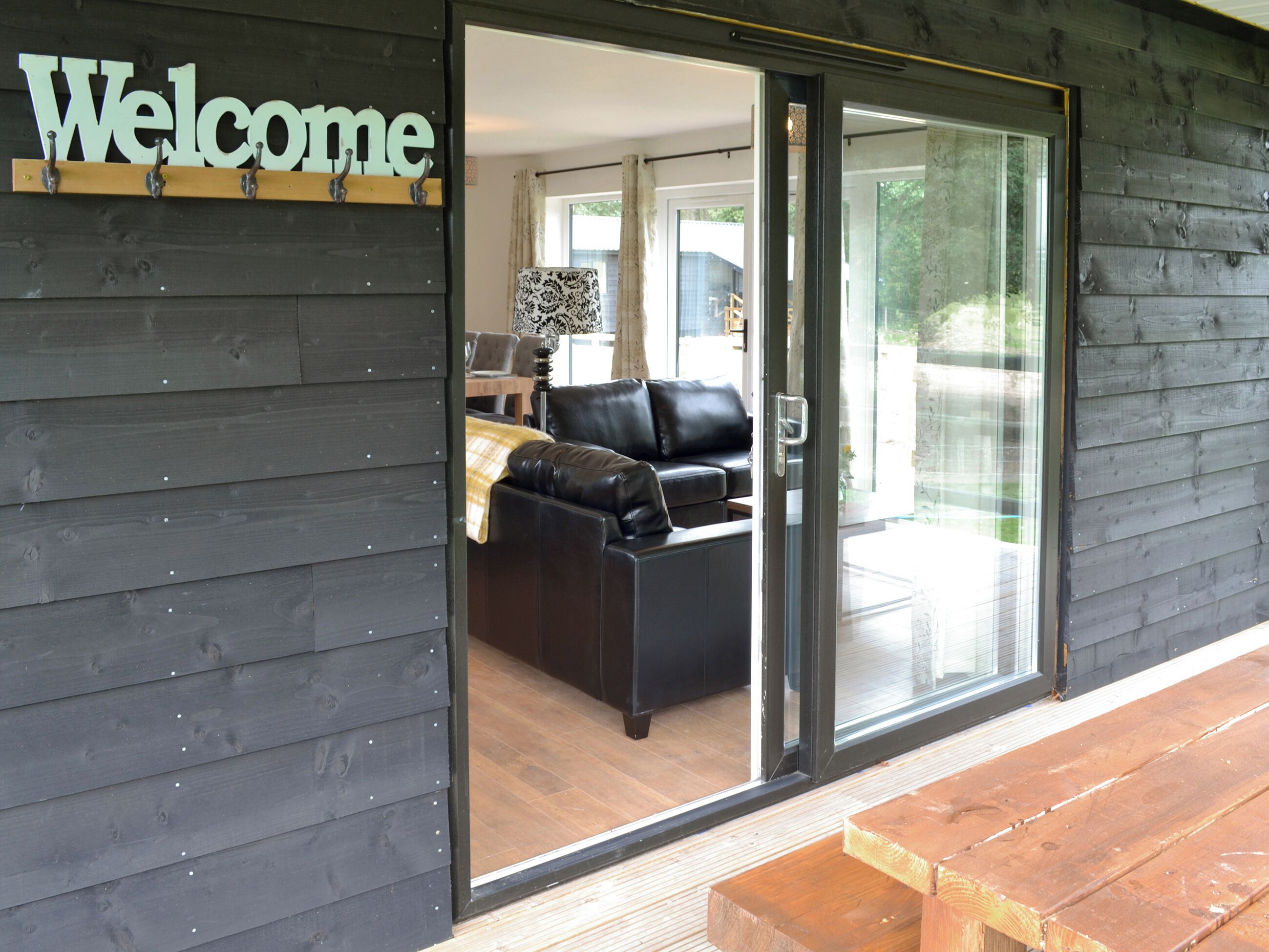 A modern building entrance with a "Welcome" sign, a sliding glass door leading to a living area with a sofa and a table.