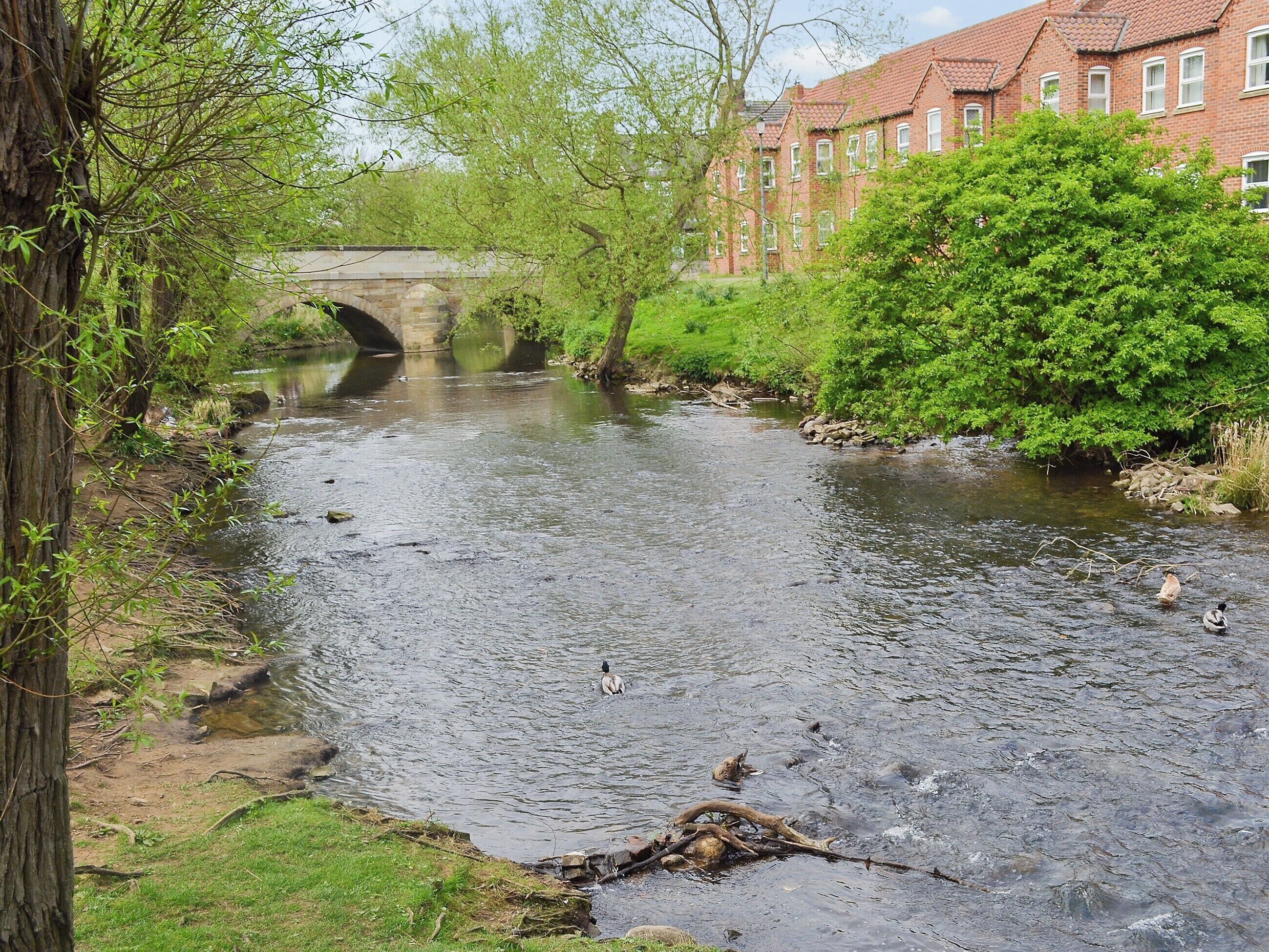 Un río con un puente de piedra, rodeado de vegetación y un edificio de ladrillo rojo.