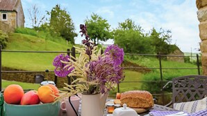 A table set outdoors with a bowl of tomatoes, a vase with purple flowers, a loaf of bread, and a plate with cutlery. - Two bedroom cottage packed with character with stone floors and wood burner. (Hawnby, near Helmsley)