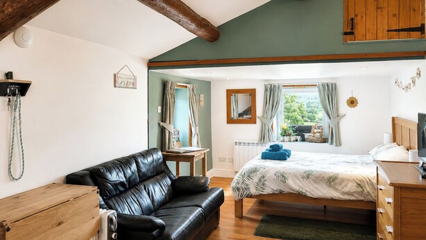 A bedroom with a bed, a black leather sofa, wooden furniture, a radiator, and a window with curtains. - The best things certainly come in small parcels and Bloomer’s Bothy has it all. (Woodhall, near Askrigg)