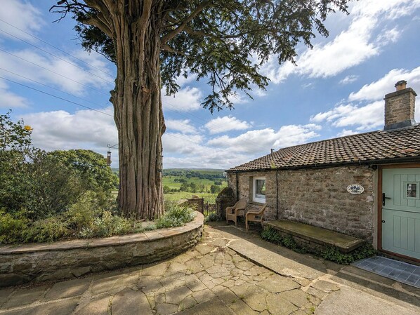 A stone building with a tiled roof, a large tree in front, and a view of the countryside. - The best things certainly come in small parcels and Bloomer’s Bothy has it all. (Woodhall, near Askrigg)