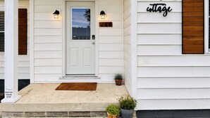 A white house with a front door, two wall-mounted lights, a wooden sign reading "lakewoods cottage", and a small potted plant on the porch. - Lakewoods Cottage (Howard)