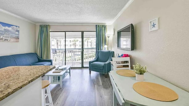 A modern living room with a blue sofa, a dining table, a TV mounted on the wall, and a balcony with curtains.