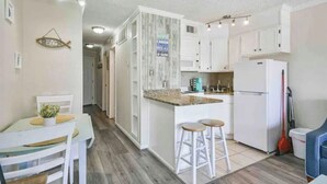 A compact kitchen with white cabinets, a granite countertop, and a built-in oven. There are two stools at the counter and a refrigerator to the right.