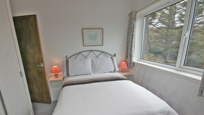 A neatly made bed with white linens, two bedside lamps, a framed picture on the wall, and a window with a view of trees.