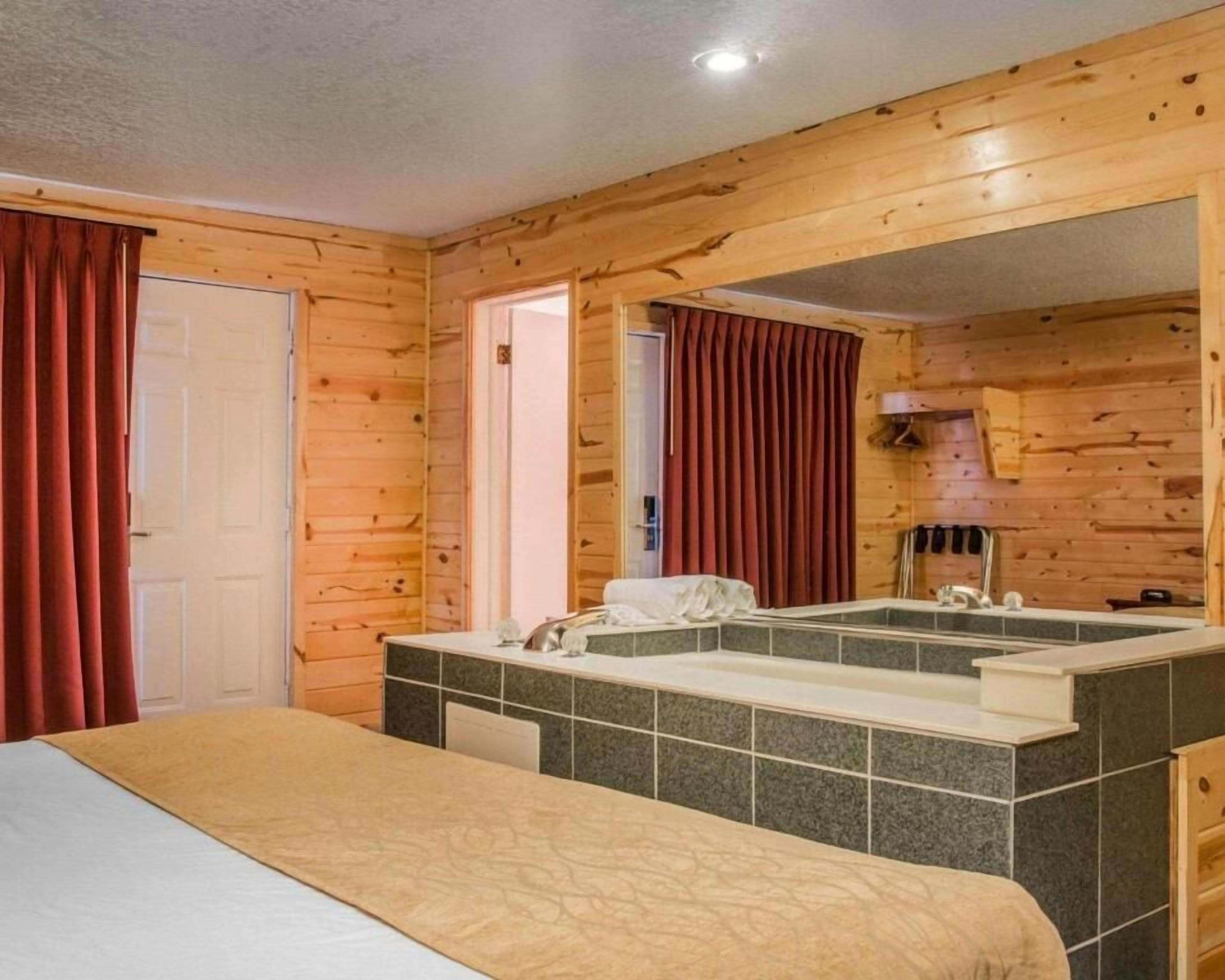 A wooden cabin room with a bed, a bathroom with a large mirror, and a window with curtains.