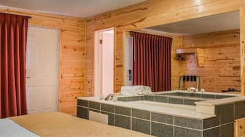 A wooden cabin room with a bed, a bathroom with a large mirror, and a window with curtains.