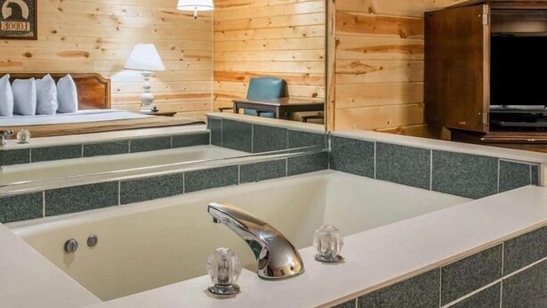 A bathroom with a large bathtub, a mirror, a sink with two faucets, and a chair. - Mackinac Lodge (Mackinaw City)