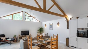 Un vaste espace de vie comprenant une table à manger en verre, des chaises en bois, un canapé, un téléviseur et une cuisine avec des meubles blancs et un four.
