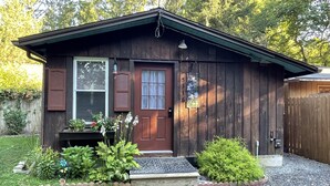 A wooden cabin with a front door, window with shutters, and a small porch area with plants.