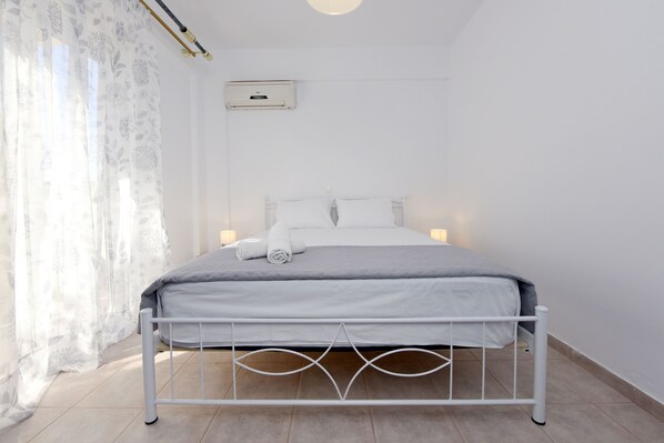 A neatly made bed with a grey blanket and white pillows, a white air conditioning unit on the wall, and a window with sheer curtains.