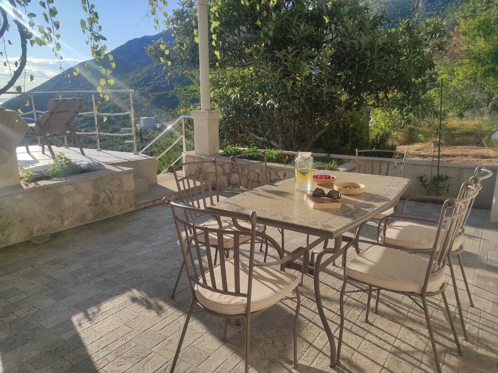 Um pátio externo com mesa e cadeiras de pedra, um pote de vidro com bebida e uma tigela de frutas.