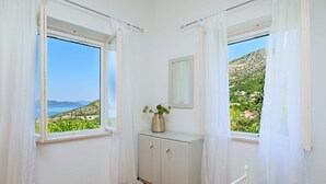 Un dormitorio con dos ventanas, una cama, una mesita de noche con un jarrón y un espejo en la pared.