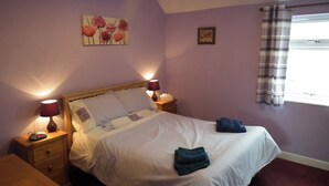 Een slaapkamer met een bed, twee nachtkastjes met lampen, een nachtkastje, een raam met gordijnen en een schilderij aan de muur.