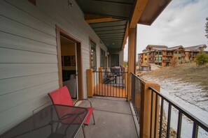 A balcony with a metal chair and a red chair, overlooking a building with multiple windows and a wooden deck. - Clearwater Lofts Bldg 3 103 (Keystone)