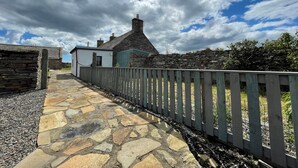 A stone pathway leading to a traditional stone house with a white exterior and a blue door, surrounded by a stone wall and a wooden fence. - You will love everything about Mey Bee. (Mey, near Thurso)
