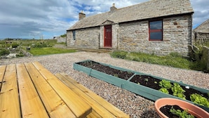 A stone cottage with a red door, a wooden bench, and raised garden beds. - You will love everything about Mey Bee. (Mey, near Thurso)