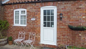 A brick house with two white-framed windows, a white door, and a small garden area with potted plants and outdoor furniture.