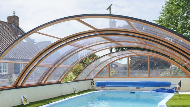 A swimming pool with a glass enclosure, surrounded by a grassy area and a brick wall.