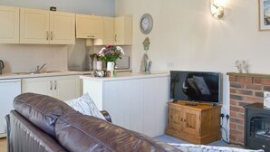 A living room with a brown leather sofa, a television on a wooden cabinet, a fireplace, and a kitchen area with white cabinets and a clock on the wall.