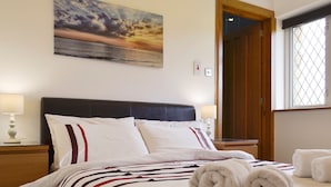 Una cama bien hecha con sábanas blancas y almohadas a rayas, un cuadro de un atardecer en la pared y una ventana con vistas al exterior.