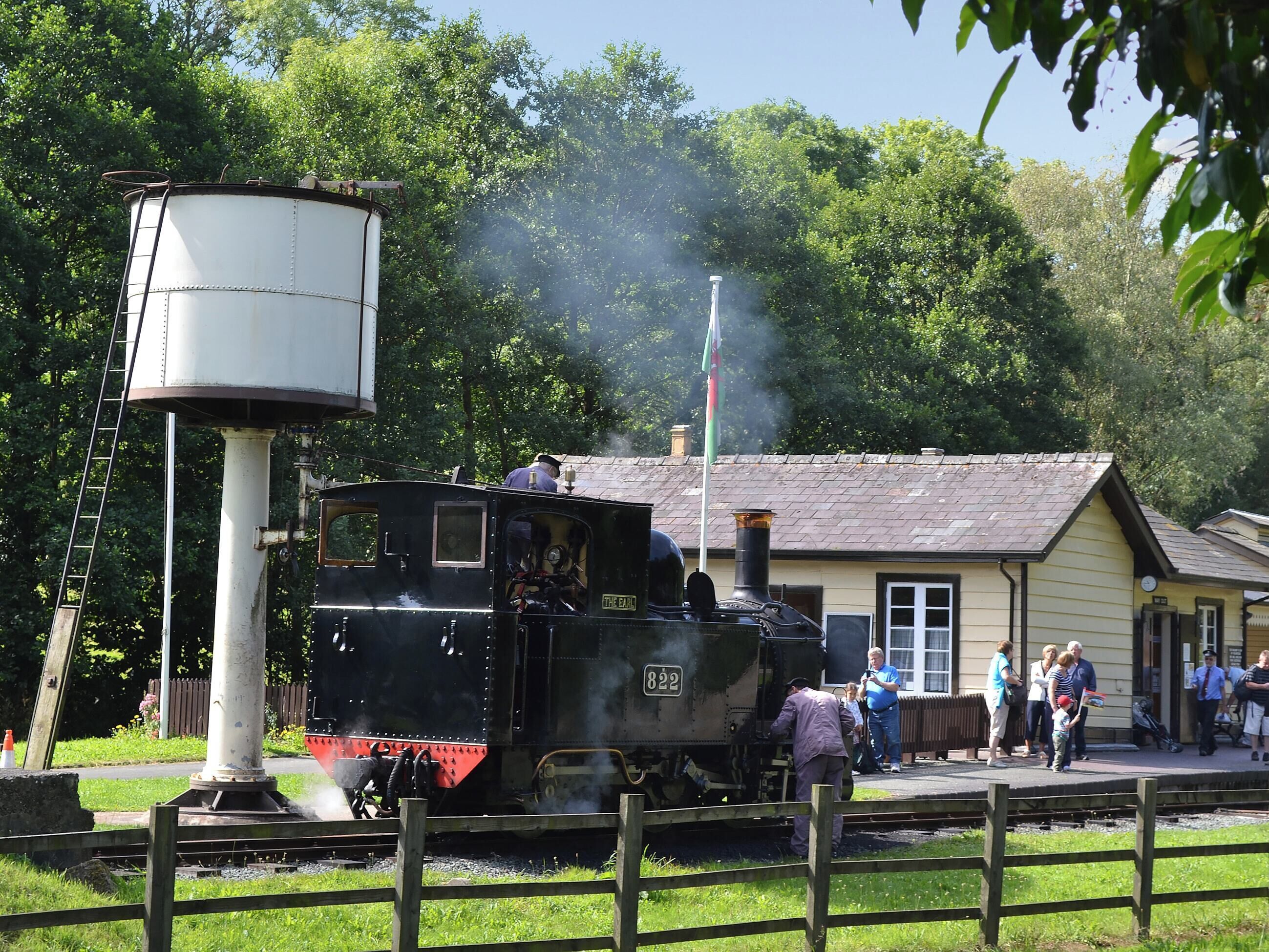 A steam locomotive at a station with a water tower, platform, and passengers.