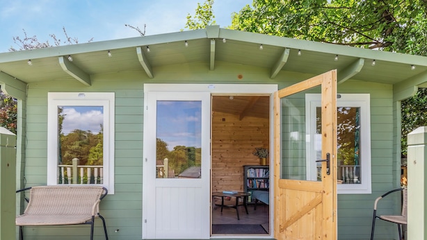 A small, green wooden building with a covered patio area, a door leading inside, and a chair outside. - 4 bedroom accommodation in Brodick (Brodick)