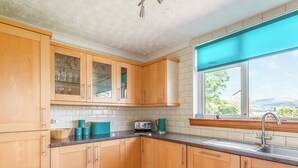 A kitchen with wooden cabinets, a window with a blue blind, and a view of greenery outside. - 4 bedroom accommodation in Brodick (Brodick)