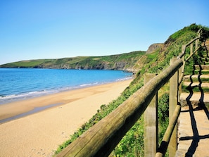 A coastal beach with a wooden fence, clear blue water, and green hills in the background.