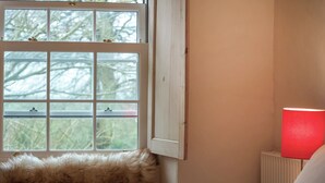 Un dormitorio con ventana, un plaid de piel, una lámpara de noche y un radiador.