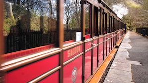 Un tren detenido, con carrocería roja y ventanas, estacionado en un andén con montañas al fondo.
