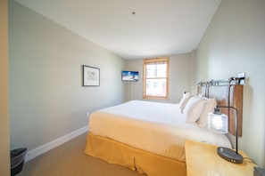 A bedroom with a bed, a nightstand, a wall-mounted lamp, a picture, and a window.