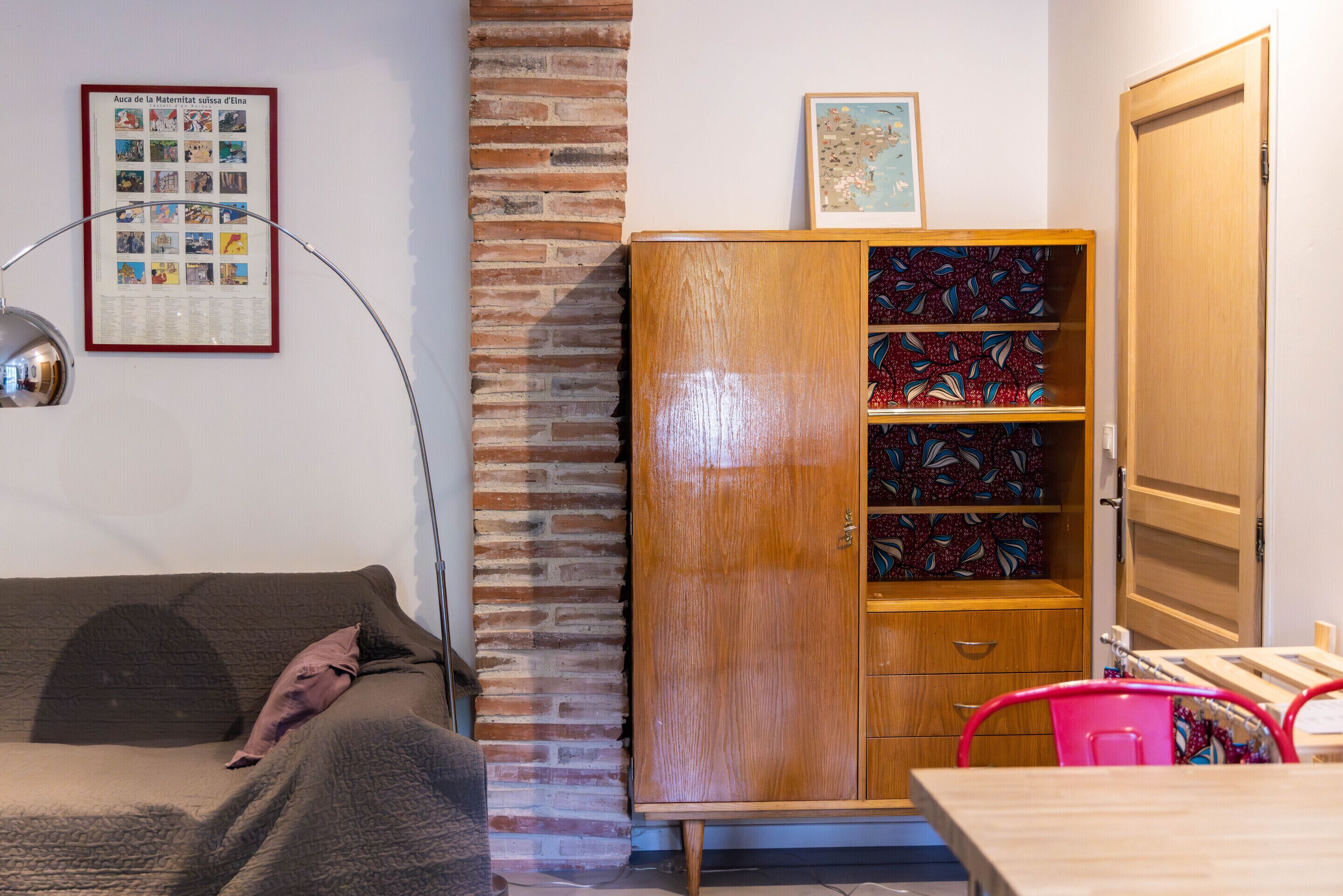 Une pièce avec un mur en briques, un meuble en bois doté de portes en verre décoré, un canapé marron, un fauteuil rose et un tableau encadré accroché au mur.