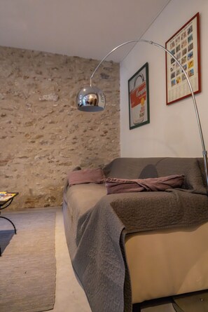 A room with a stone wall, a bed with a blanket, a floor lamp, and a poster on the wall.