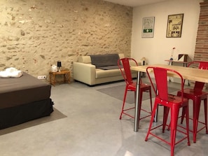 Una habitación con pared de piedra, un sofá, una mesa de comedor con sillas rojas y una mesita auxiliar con una lámpara.