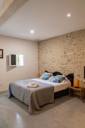 A bedroom with a stone wall, a bed with a grey bedspread, two pillows, a bedside table with a lamp, and a window with a view.