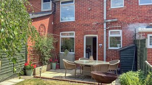A backyard with a brick house, a round table with chairs, a barbecue grill, and potted plants. - 2 bedroom accommodation in Stockbridge (Stockbridge)