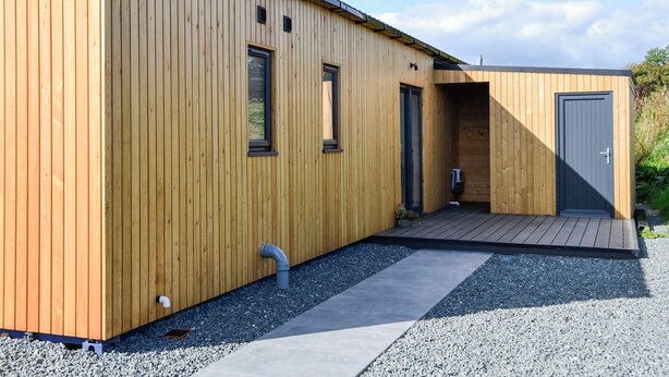 A modern wooden building with a flat roof, two windows, a door, and a small outdoor area with a pathway.