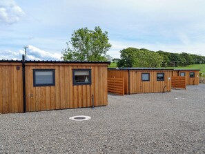 A row of wooden cabins with individual windows and doors, arranged on a gravel surface with a clear sky and trees in the background.