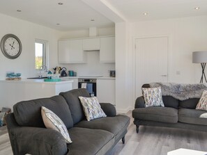 A modern living room with two grey sofas, a kitchen with white cabinets, and a clock on the wall.