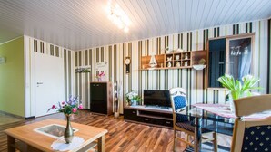 A modern living room with striped wallpaper, a wooden entertainment unit, a flat-screen TV, a dining table with chairs, and a small kitchen area.