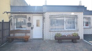A building with a white brick facade, a black roof, and a white door. There are two windows with decorative iron grilles, and a bench is placed outside.