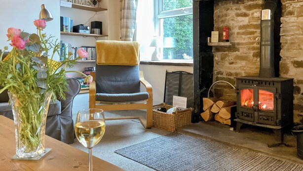 A cozy living room with a fireplace, a comfortable chair, a table with a vase of flowers and a glass of wine, and a bookshelf.